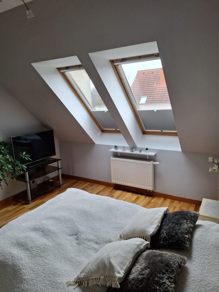 Plisy na okna dachowe w sypialni – realizacja Veo Design Łódź Sypialnia na poddaszu z dwoma oknami dachowymi wyposażonymi w plisy w kolorze jasnoszarym. Wnętrze z białą pościelą, dekoracyjnymi poduszkami i drewnianą podłogą.